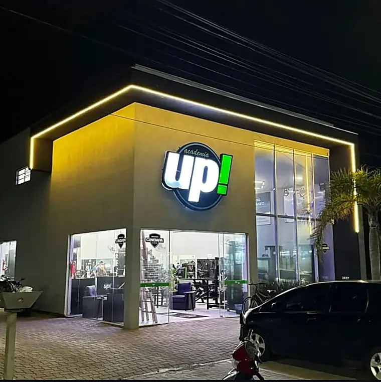 Fachada da Academia Up! à noite, em Imbé: fita de LED contornando o beiral, letreiro retroiluminado e vidro mostrando o salão de treino.